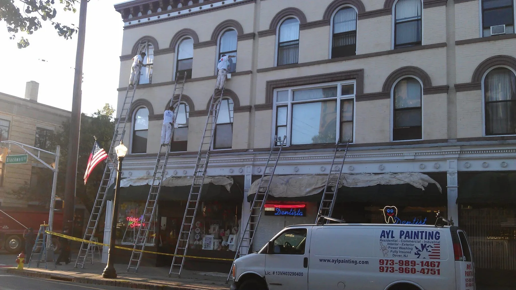 Three A.Y.L. painters on extension ladders painting the upper facade of a commercial building with storefronts at street level.