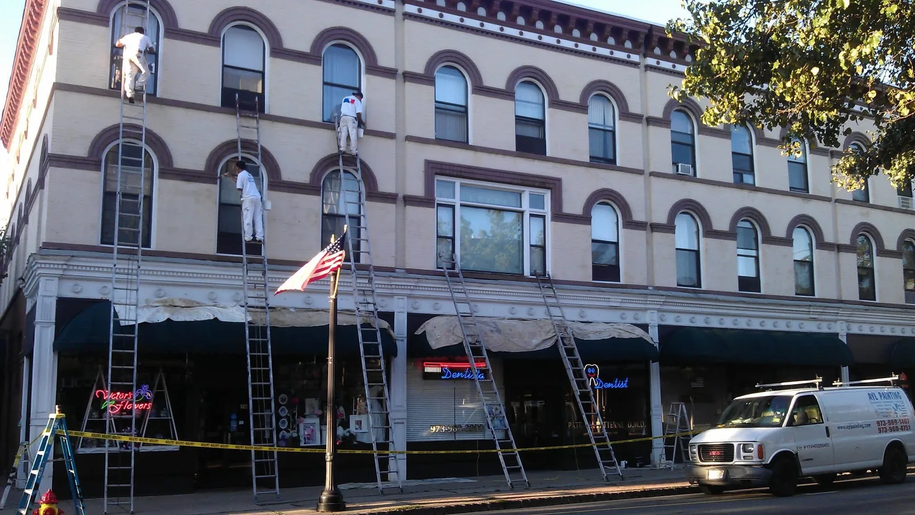 Four painters on extension ladders working across the three-storey facade of a historic downtown building above its storefronts.