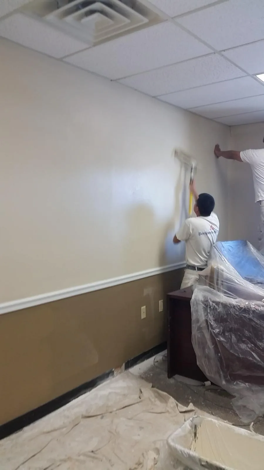 Two painters rolling and cutting in the walls of a commercial interior with furniture covered in plastic.