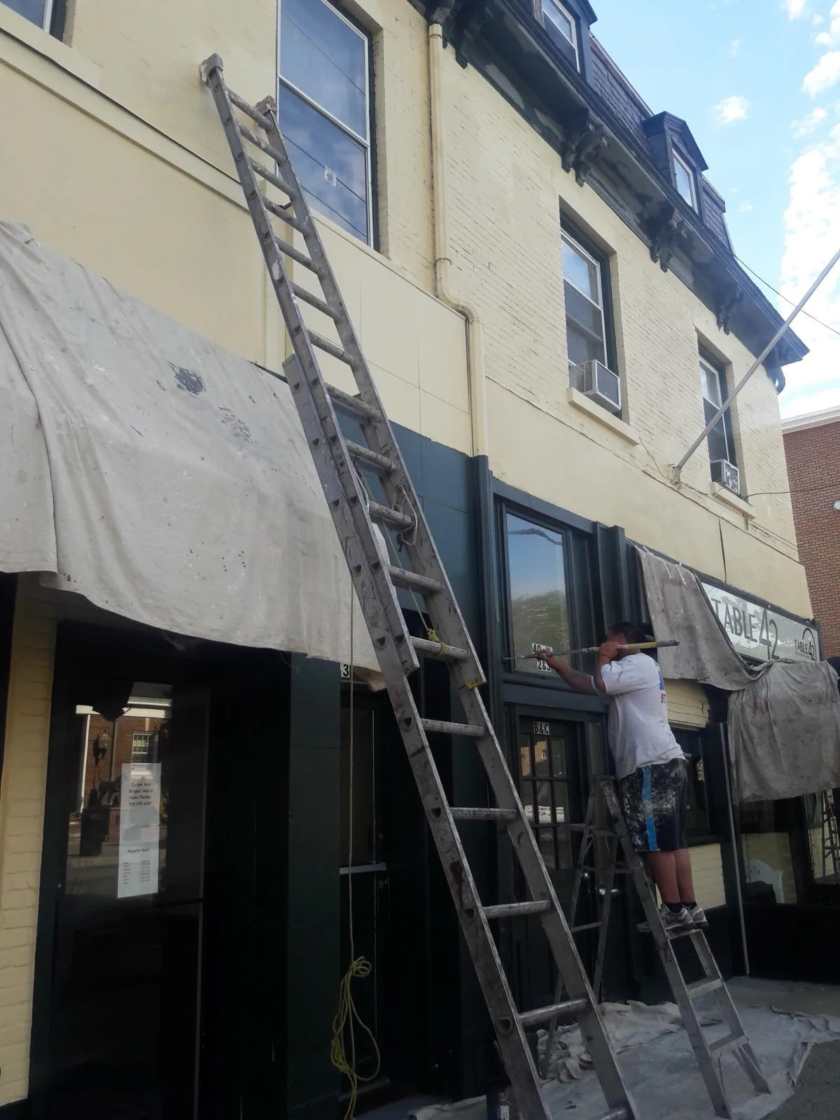 A painter on a ladder cutting in trim above a storefront, with awnings and windows masked off with drop cloths.
