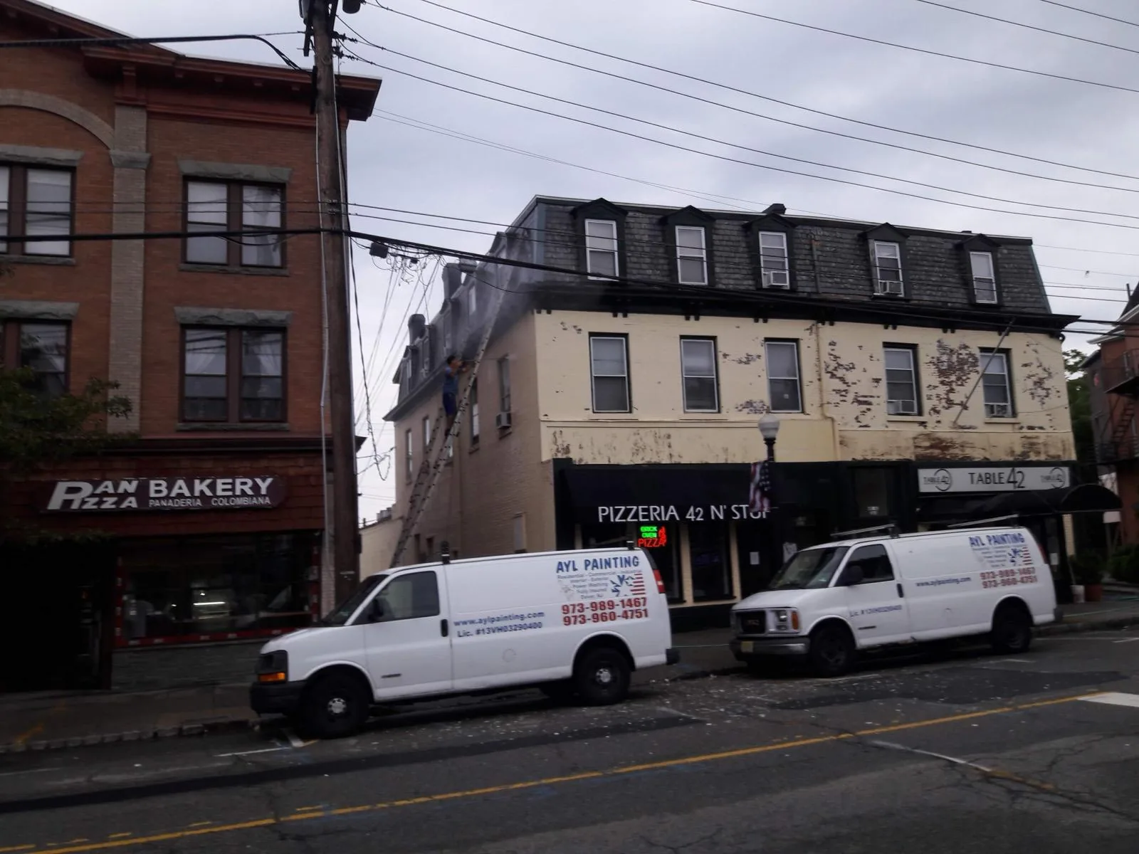 Two A.Y.L. Painting vans parked along a street of storefronts while a painter works from a ladder on the peeling upper facade.