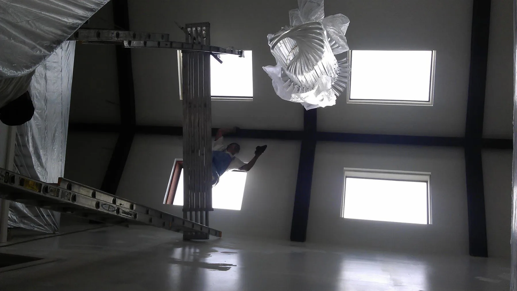A painter working at height on the ceiling of a large open interior with fixtures covered in plastic.