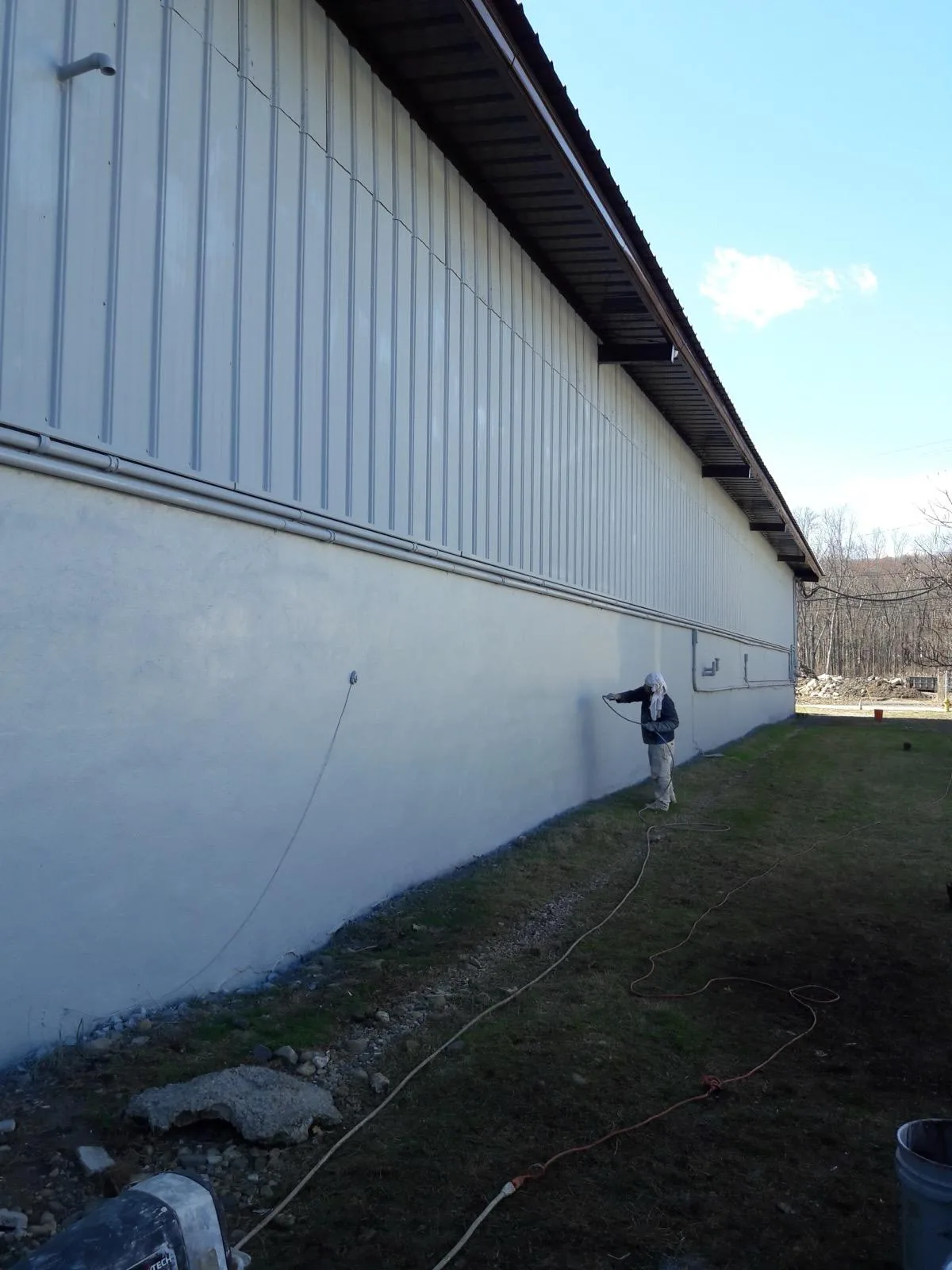 A painter spraying the exterior wall of a long metal-sided industrial building.