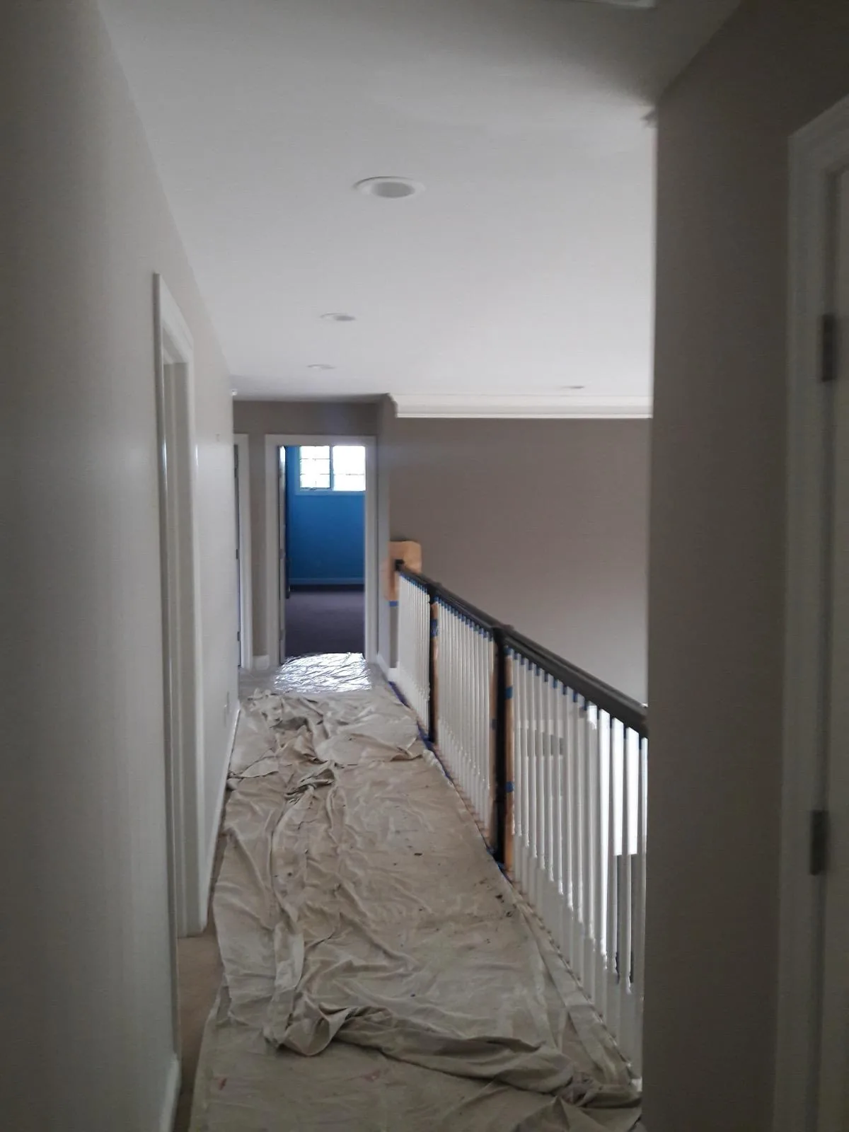 An upstairs hallway with freshly painted walls and ceiling, drop cloths along the floor and the stair railing masked with tape.