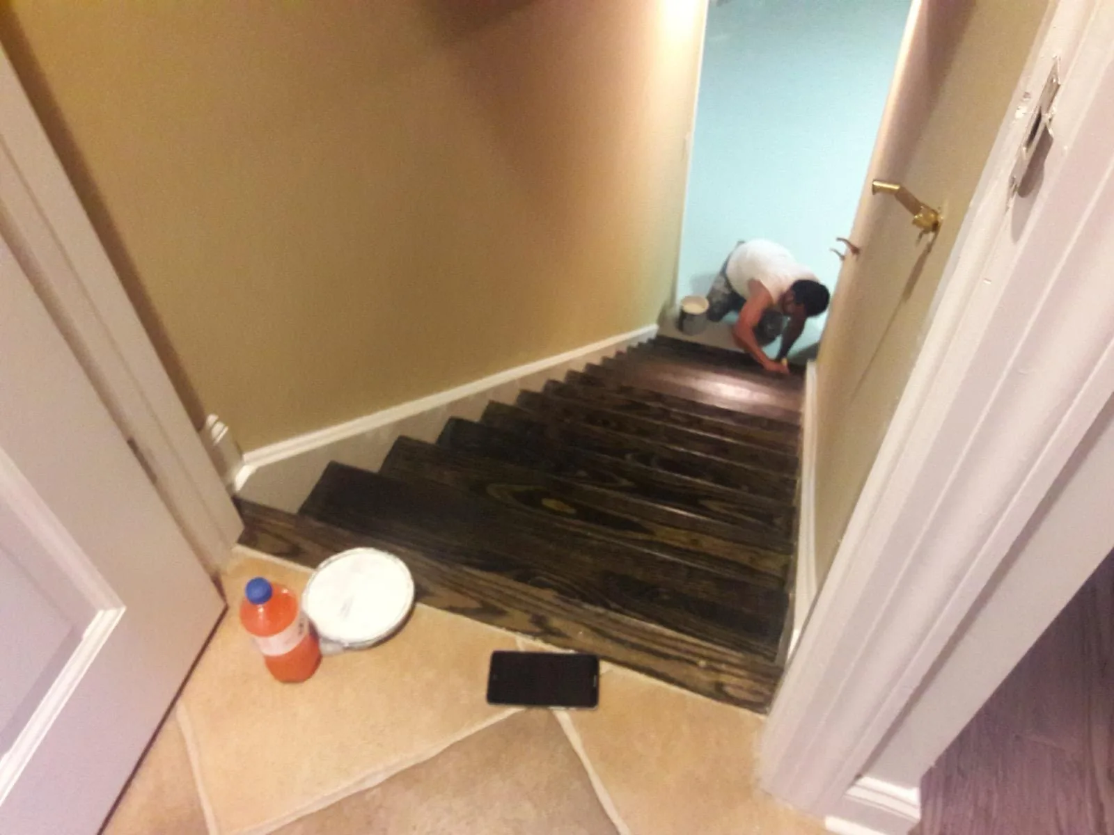 A painter finishing dark-stained stair treads at the top of a staircase, with a paint can and cut-in brush at hand.