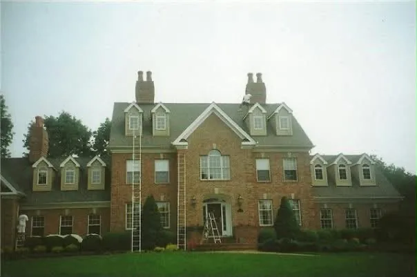 A large brick house with multiple dormers and chimneys, ladders set against the front elevation and painters at work.