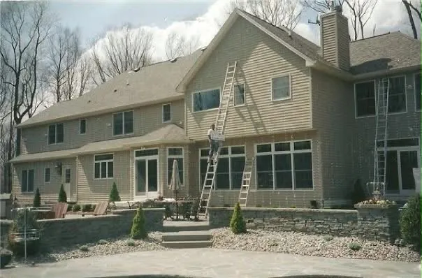 The long rear elevation of a large house with two extension ladders in place and a painter working on the upper siding.