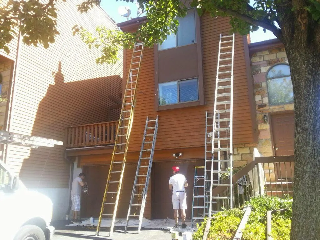 Painters working from ladders on the exterior siding and garage doors of brown-sided multi-unit homes.