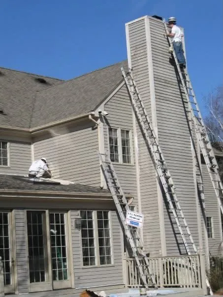 A painter on a tall extension ladder working on the chimney of a grey-sided house while a second painter works on the lower roof.