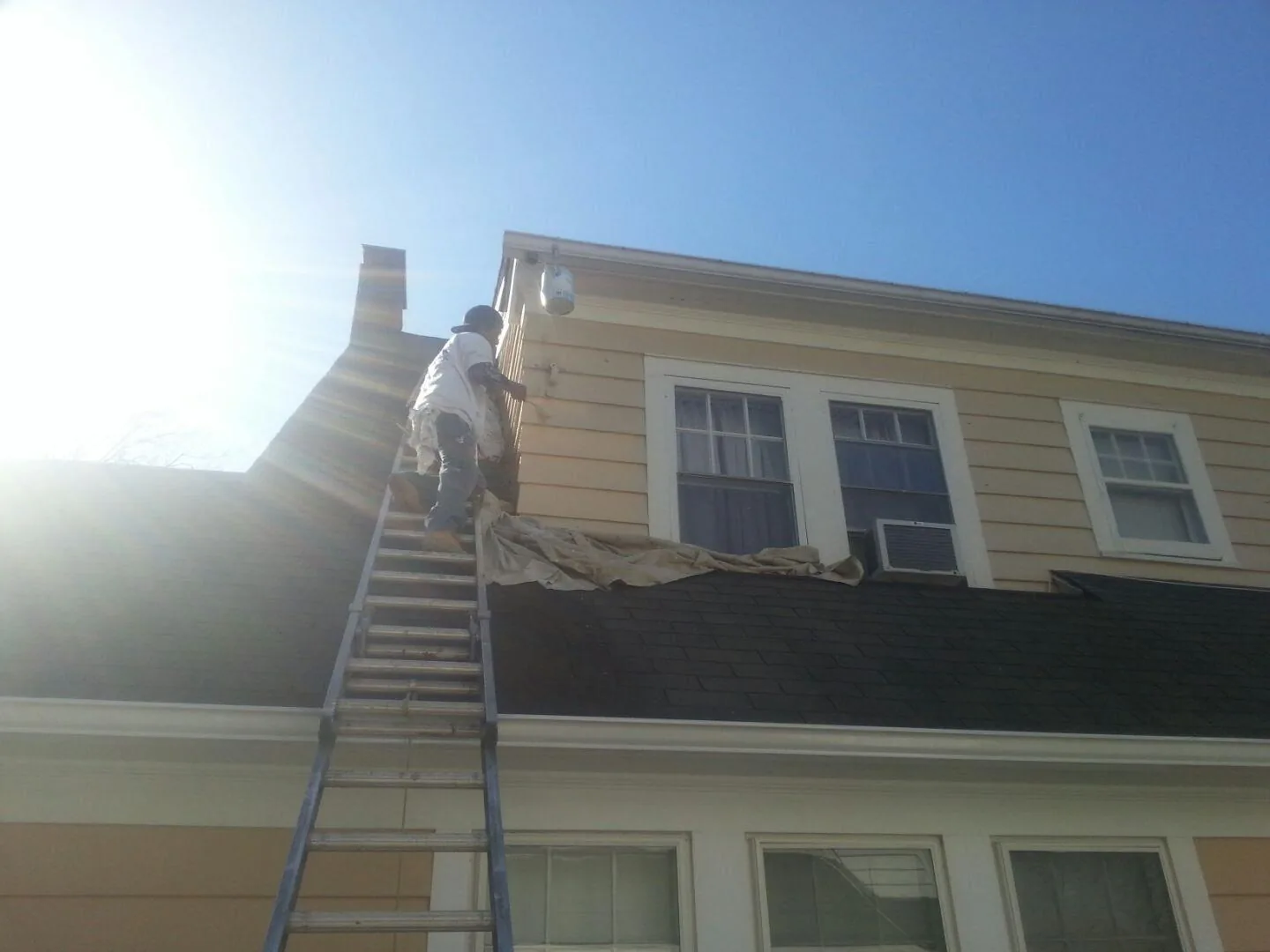 A painter high on a ladder working on second-storey siding beside a chimney, with a drop cloth spread over the lower roof.