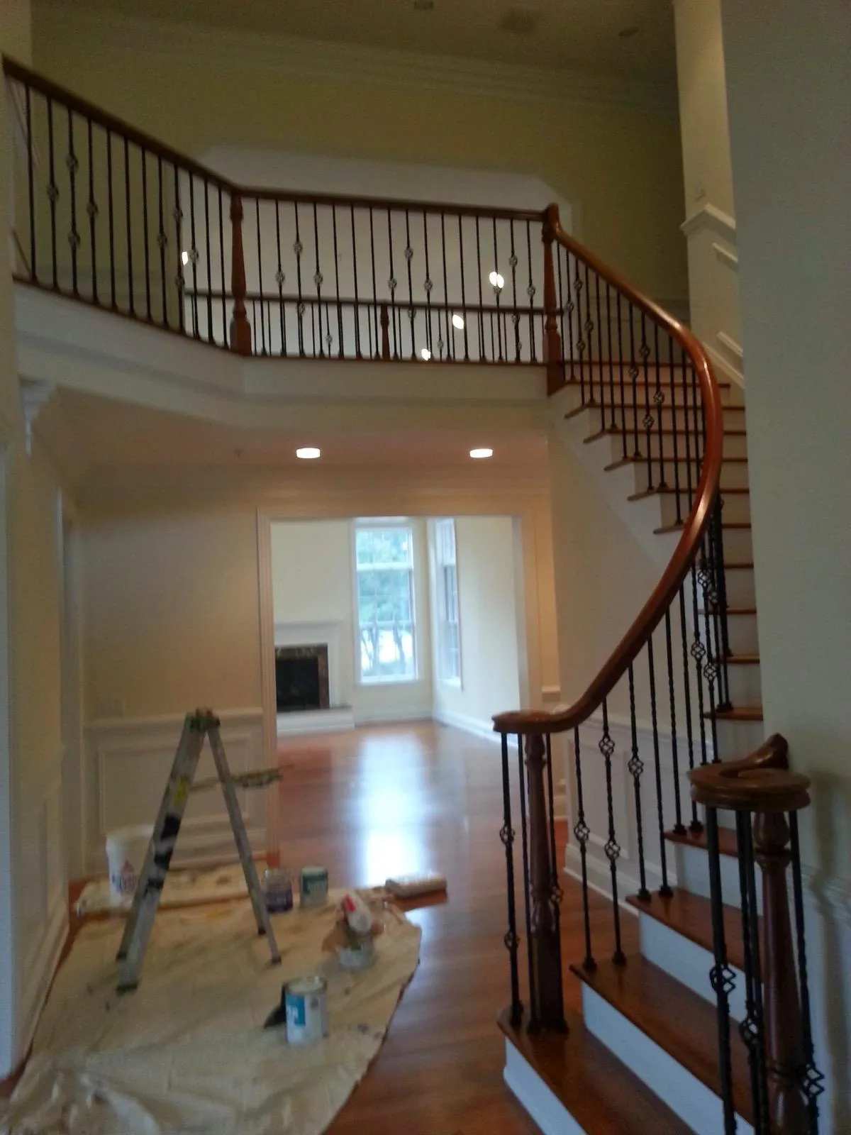 A two-storey foyer with a curved staircase, wood handrail and white wainscoting, with drop cloths and a ladder in place.