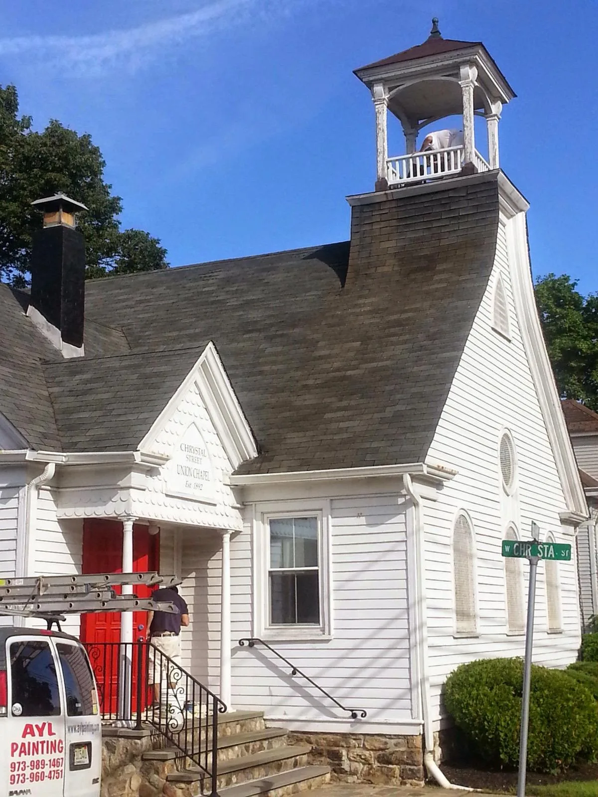 A painter working in the open bell tower of a white clapboard chapel.