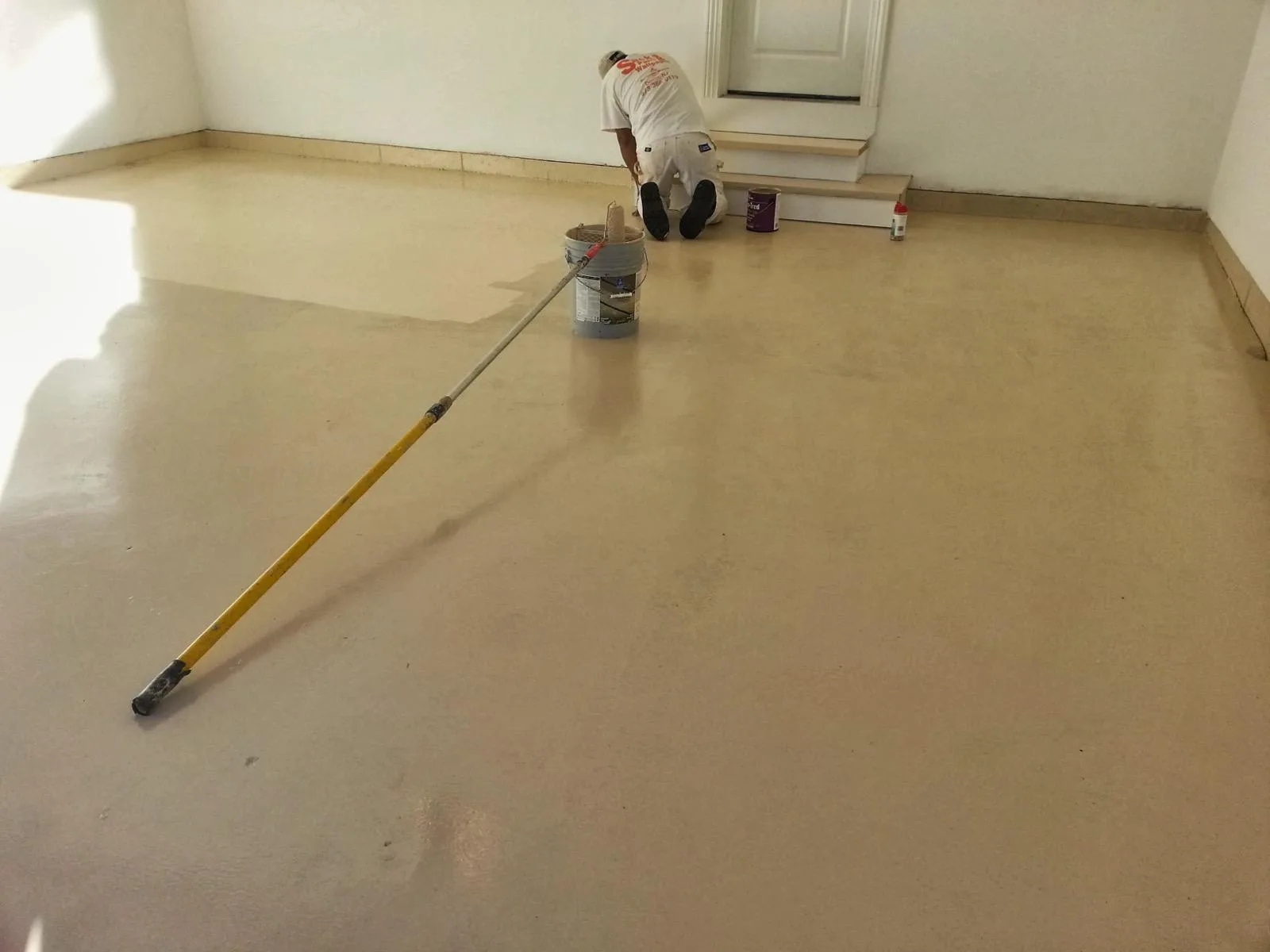 A painter applying floor coating across a large interior floor with a roller on an extension pole.