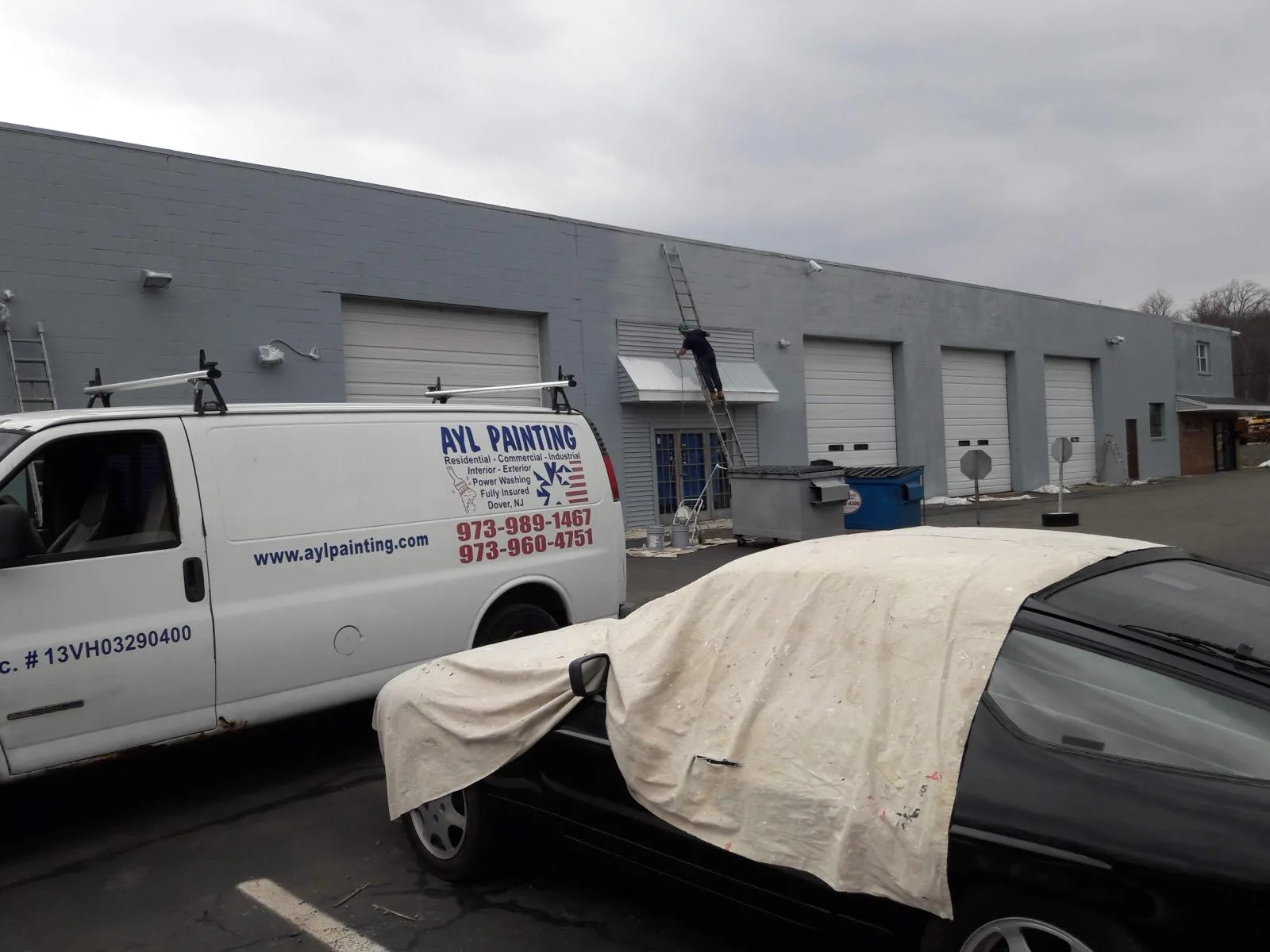 An A.Y.L. Painting van outside a grey industrial building with roll-up doors while a painter works from a ladder above the entrance.